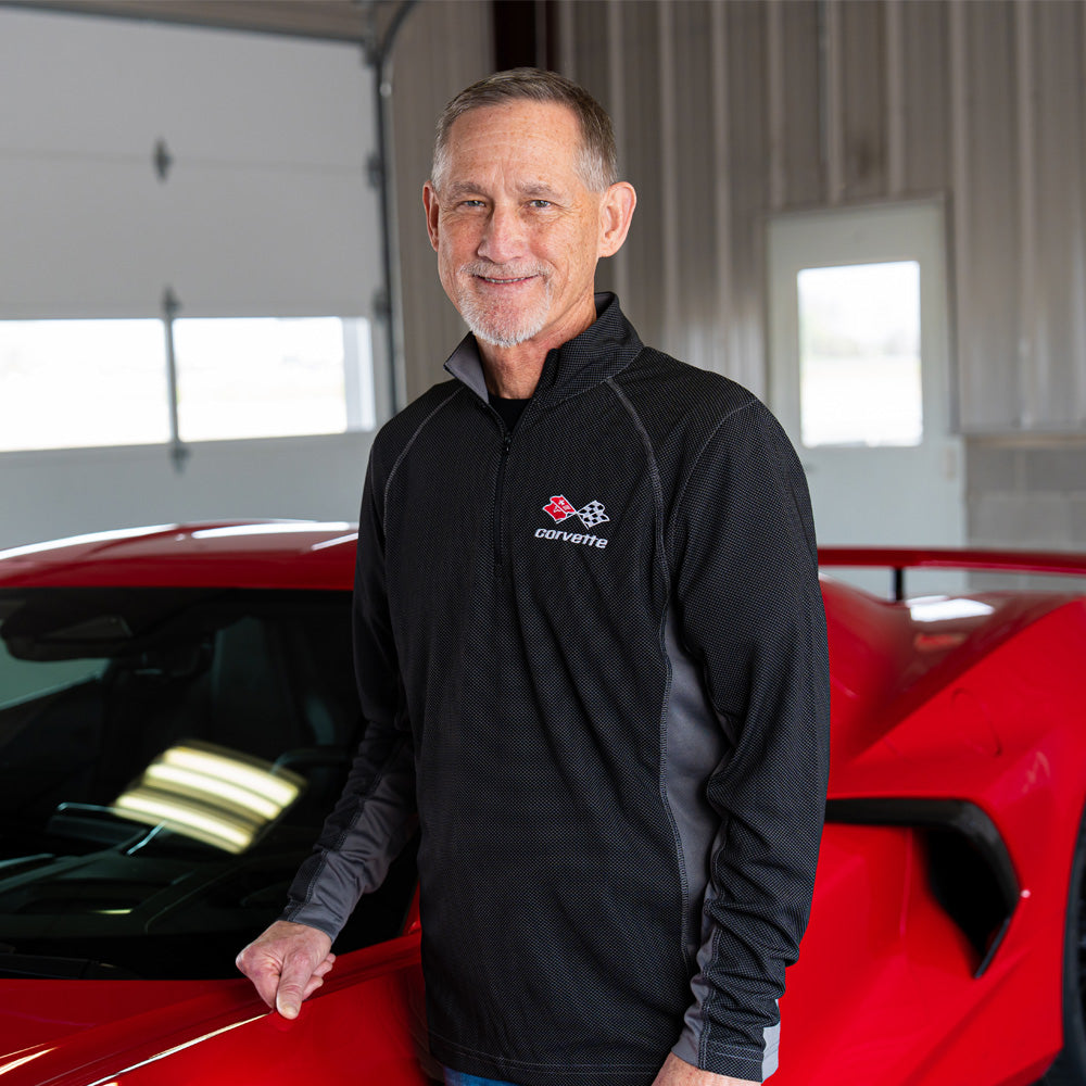 Man wearing the 3rd Generation Corvette Emblem Ice Colorblock Quarter-Zip Pullover in black and gray standing next to a red Corvette.