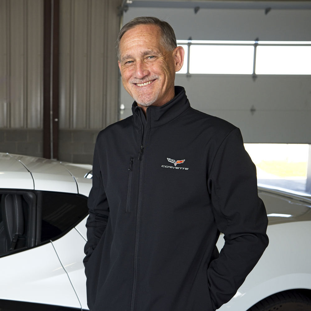 Man standing in front of a white Corvette wearing a black softshell jacket with a C6 Corvette emblem embroidered on the left chest.
