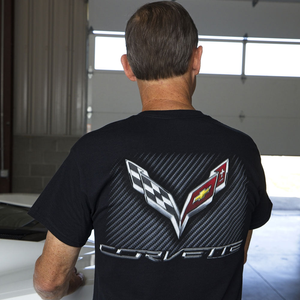 A man standing in a garage showing the large C7 Corvette emblem on a carbon fiber background on the back of a black t-shirt.