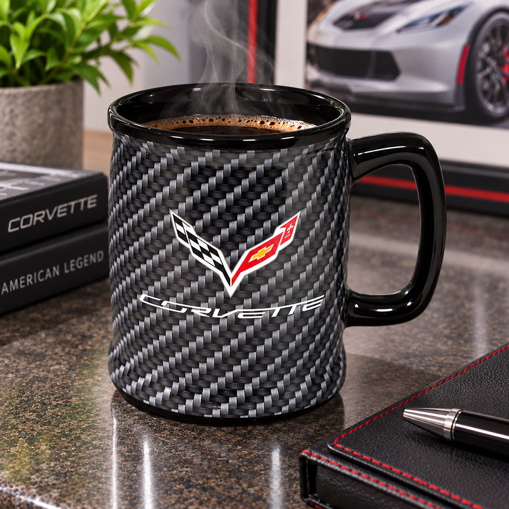 A black ceramic coffee mug with a carbon fiber pattern and a C7 Corvette logo, filled with steaming coffee on a desk.