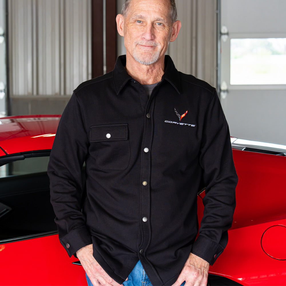 Man wearing a black Corvette shirt standing next to a red Corvette in a garage.