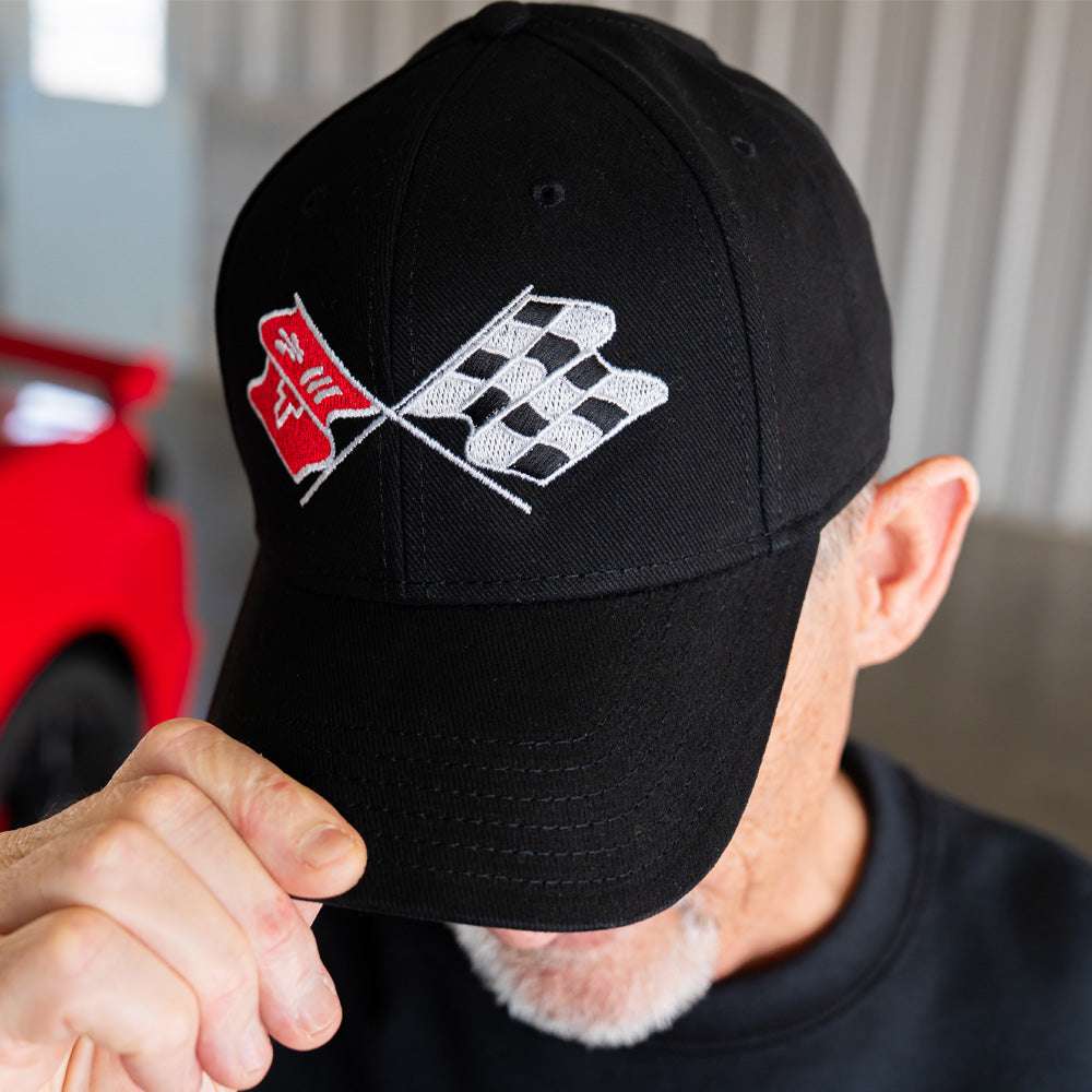 Person wearing a black cap with an embroidered C3 Corvette emblem, with a blurred red Corvette in the background.