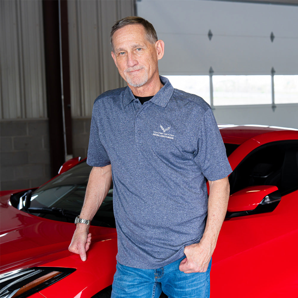 Man in a heather blue C7 Corvette polo shirt standing next to a red Corvette in a garage