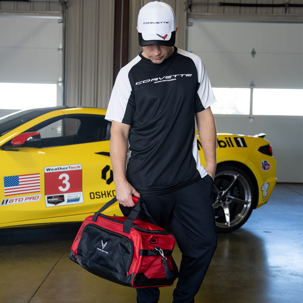 Person holding a Corvette red duffel bag in front of a yellow Corvette race car in a garage.