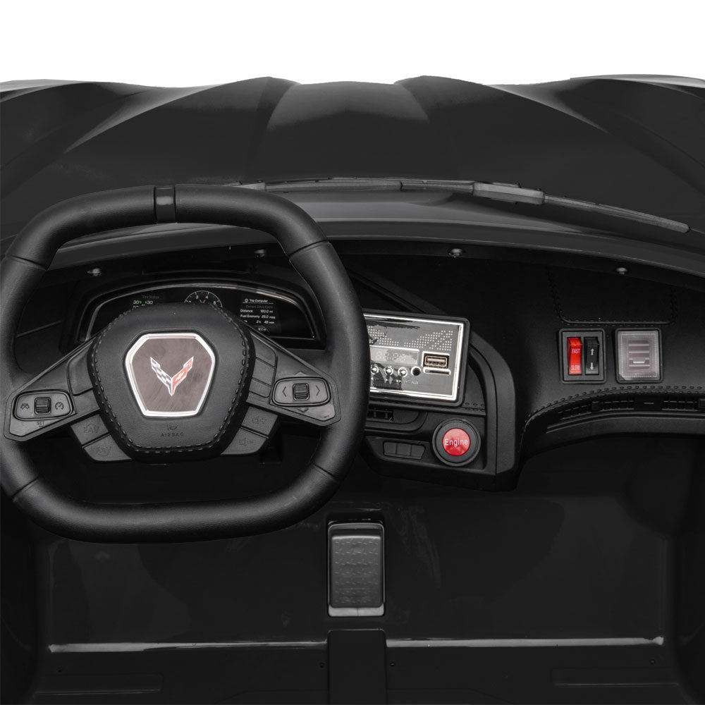 Close-up of a black convertible ride-on car dashboard and steering wheel, featuring a C8 Corvette emblem on the wheel center, a small screen, and red and silver control buttons.