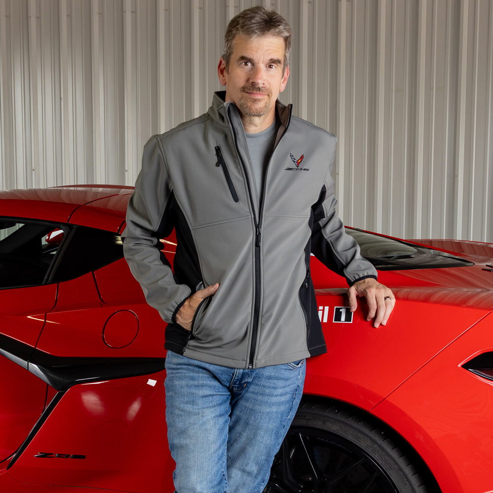 Man wearing a gray and black colorblock jacket with the C8 Z06 emblem on the left chest while standing next to a red Z06 Corvette.
