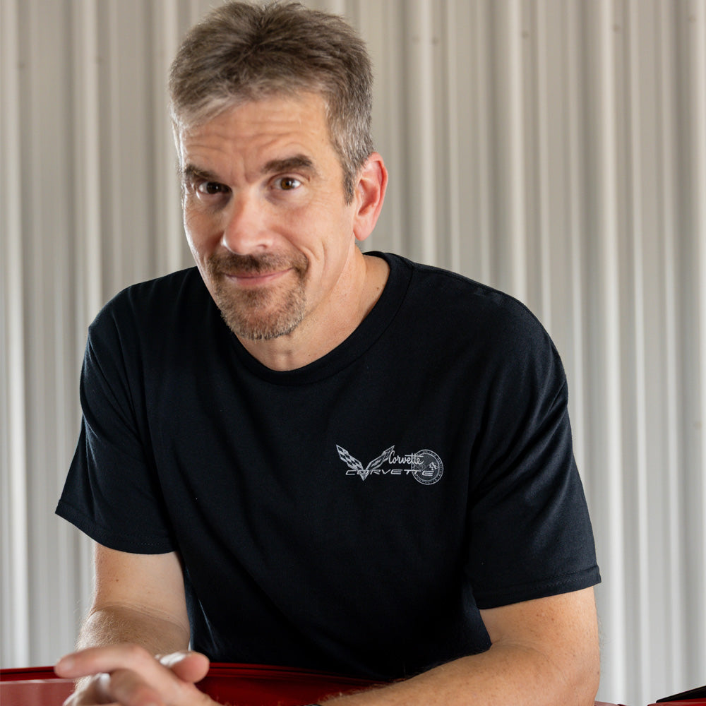 Man wearing a black Corvette t-shirt with a small Corvette logo on the left chest, sitting against a neutral background