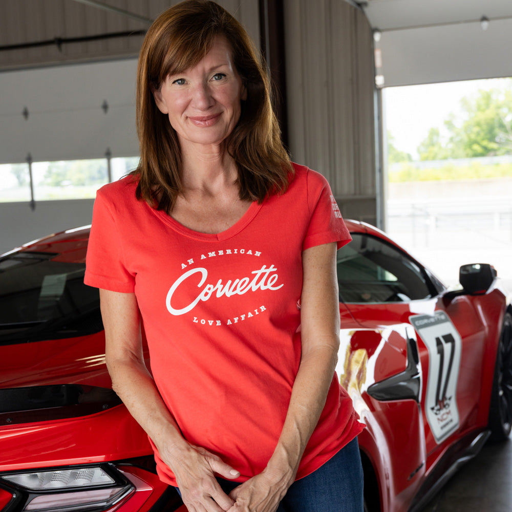 Woman wearing a red Corvette t-shirt standing in front of a red Corvette car in a garage.