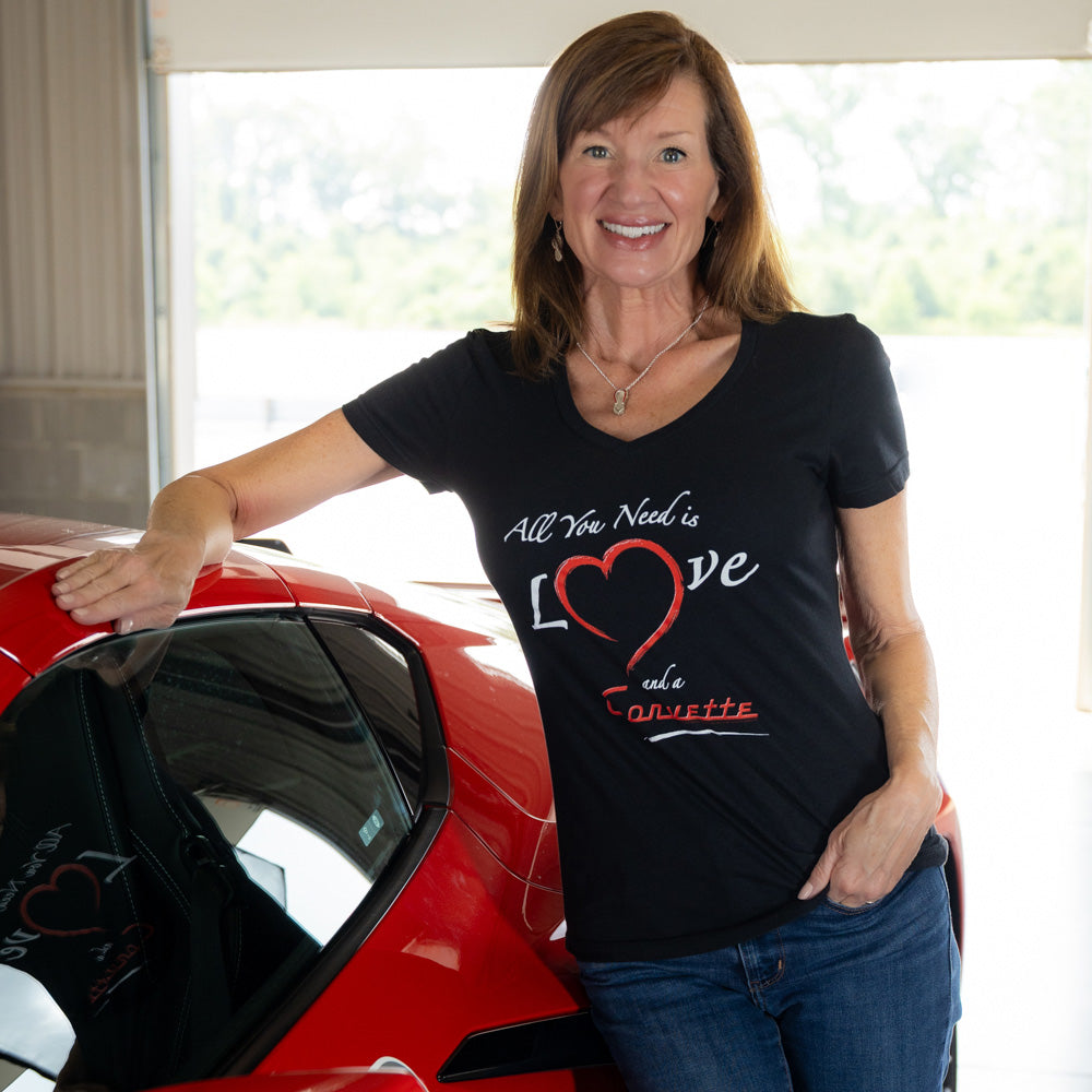 Woman wearing a black t-shirt with a All you need is love and aCorvette graphic, standing next to a red Corvette.
