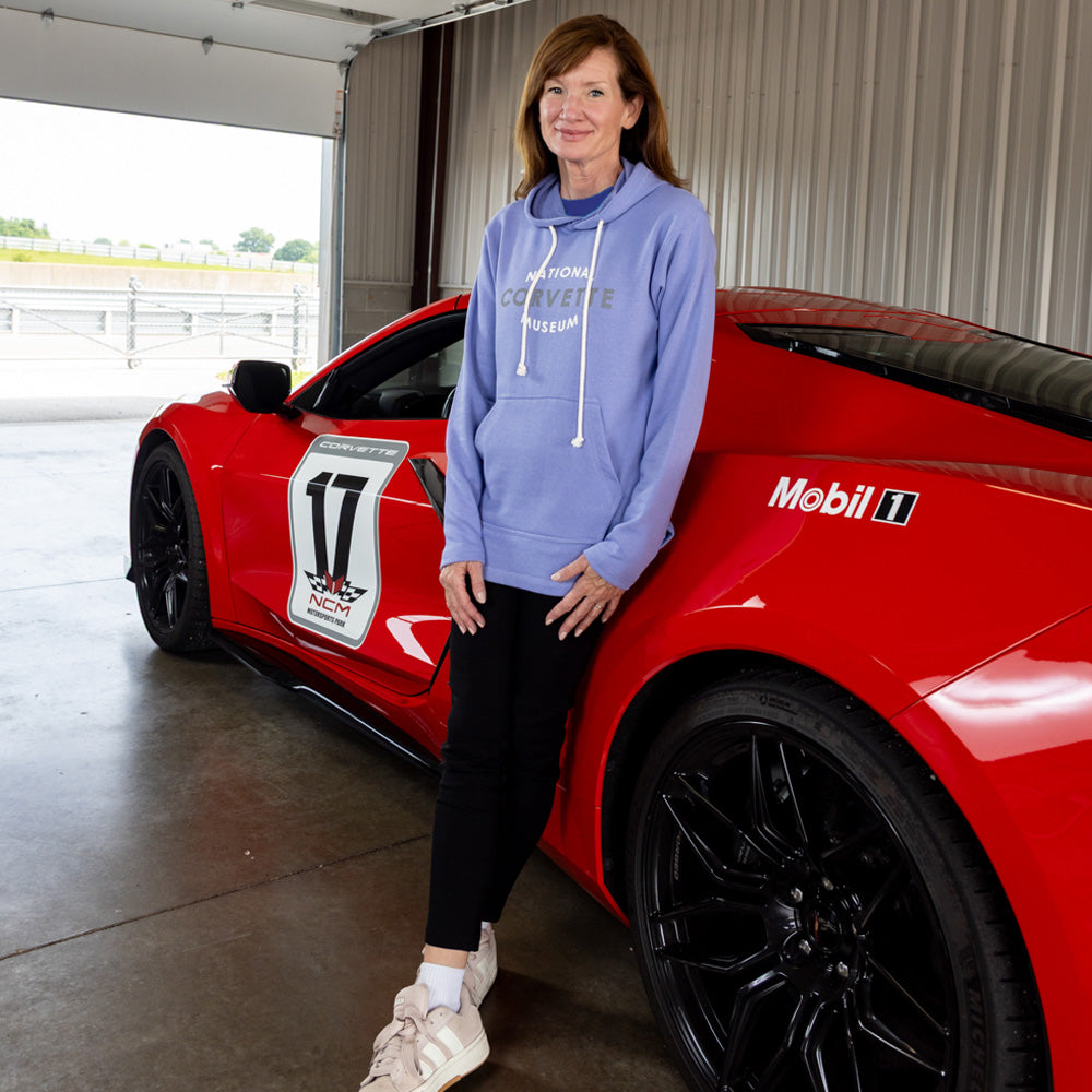 Woman in a purple National Corvette Museum hoodie standing next to a red Corvette in a garage.