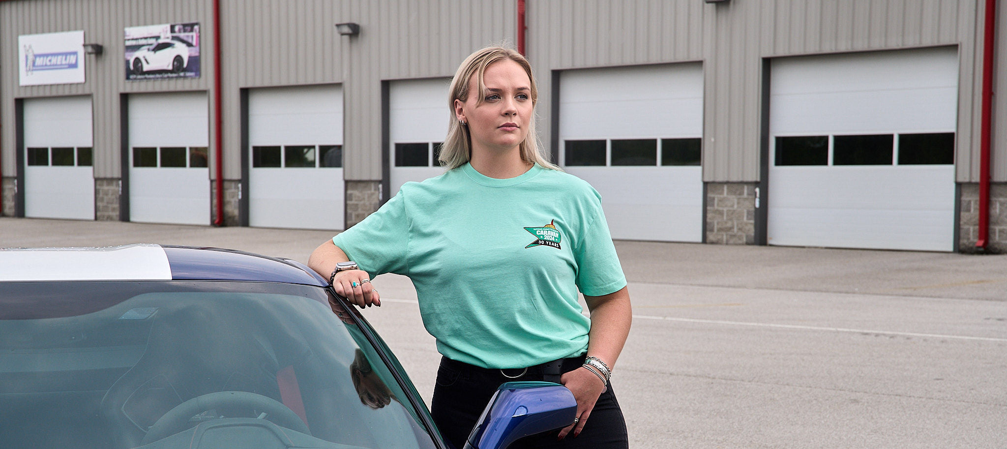 blonde female wearing teal shirt standing next to Corvette