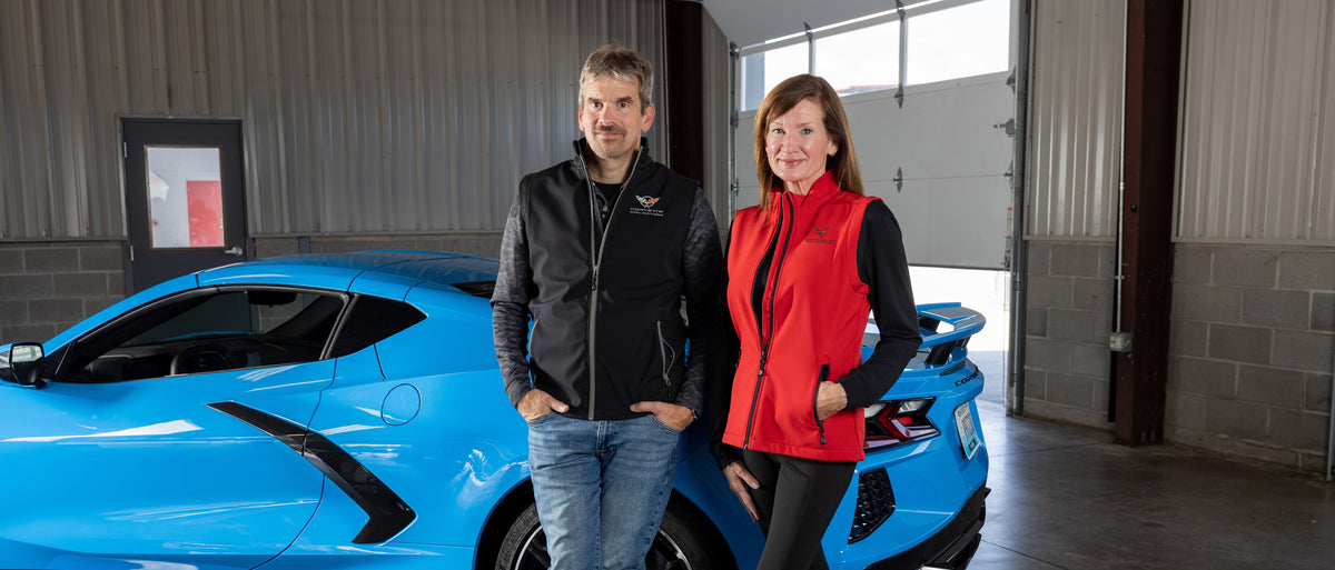 A man and woman each wearing a vest with an embroidered Corvette emblem on the left chest and standing in front of a blue Corvette