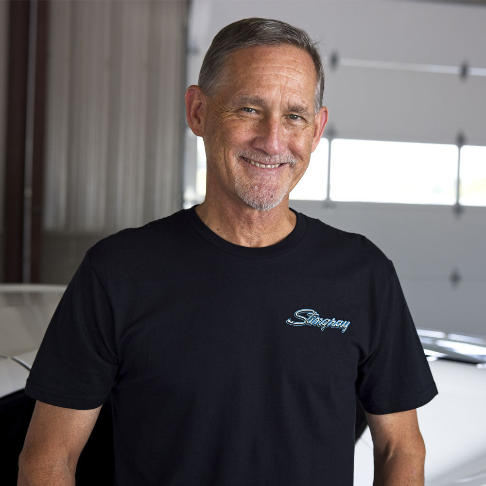 Man standing in a garage next to a white Corvette wearing a black t-shirt with the word Stingray on the left chest.