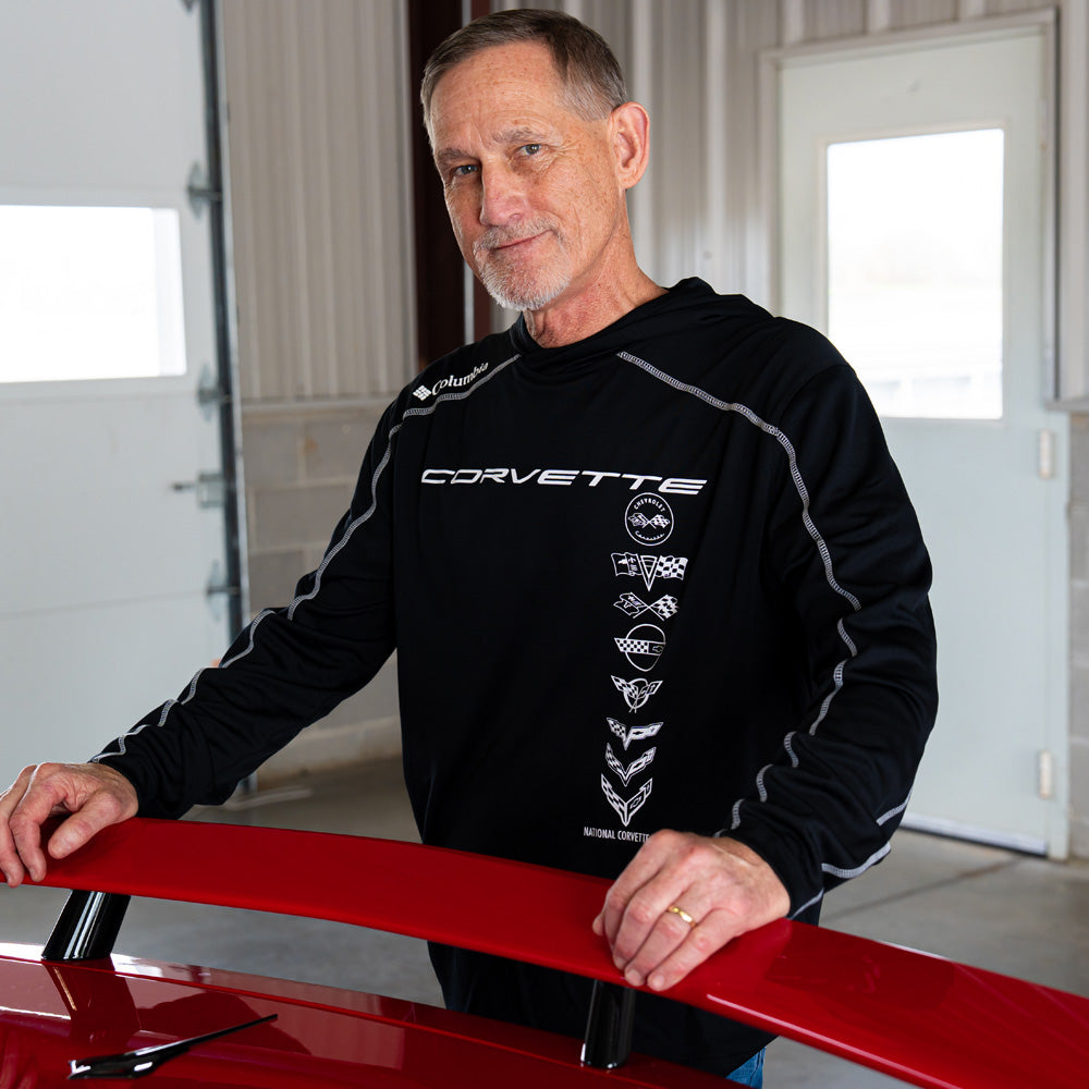 Man wearing a black Corvette Columbia pullover standing next to a red Corvette in a garage.