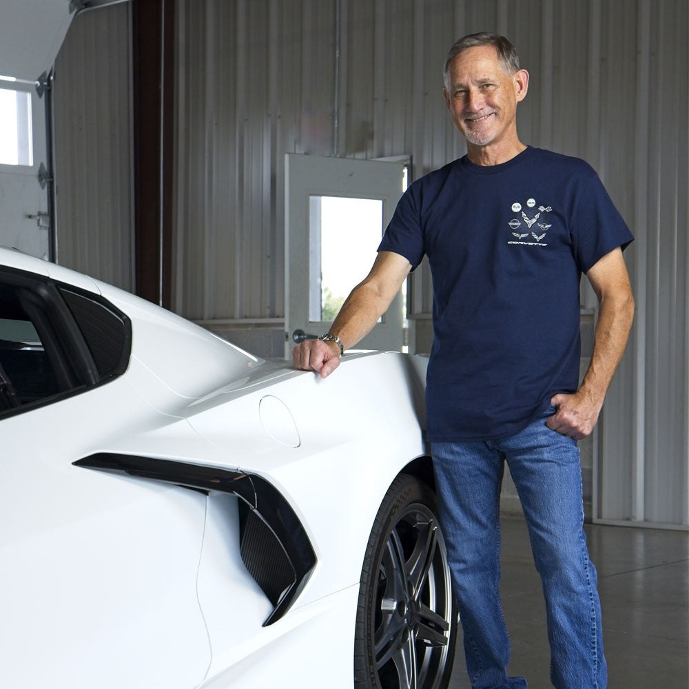 A man standing in a garage next to a white Corvette wearing a navy tee showing a C1-C8 emblem design on the front left of the tee.