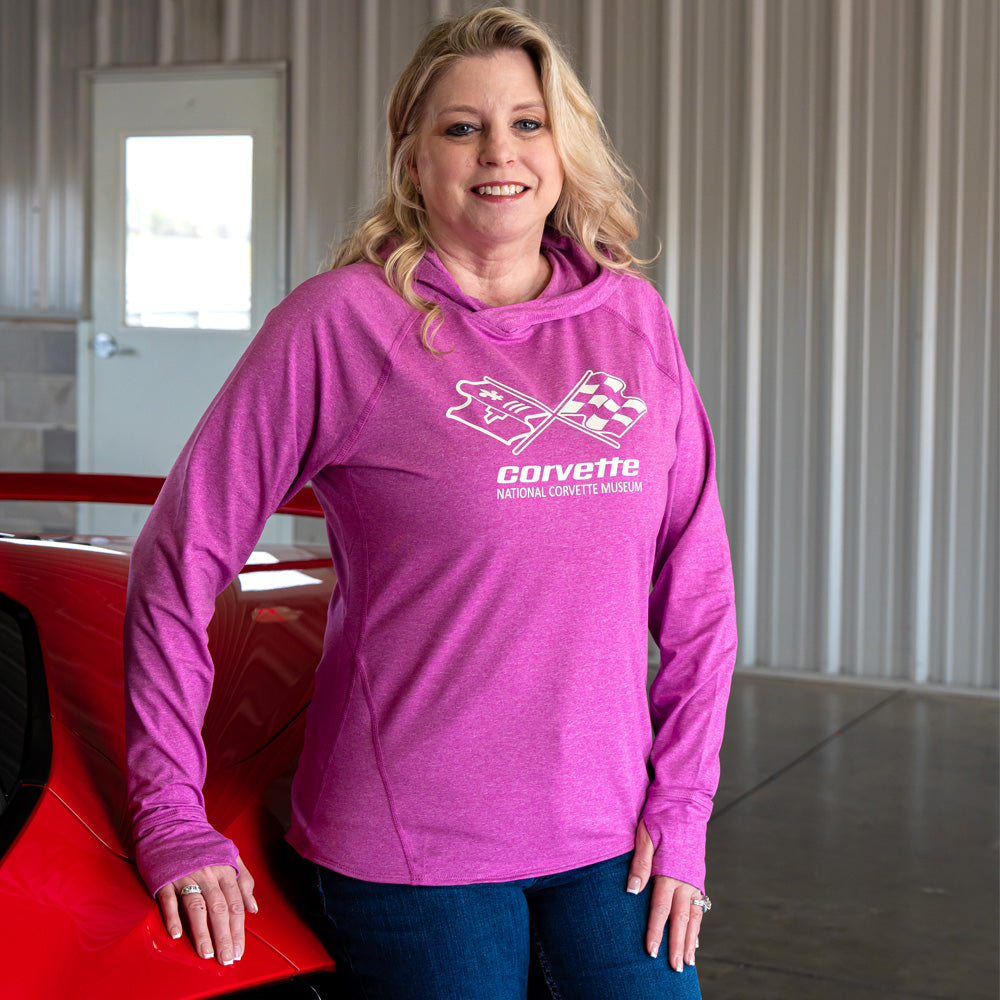 Woman wearing a hot pink pullover with the C3 Corvette emblem design on the front chest standing next to a red Corvette parked in a garage.