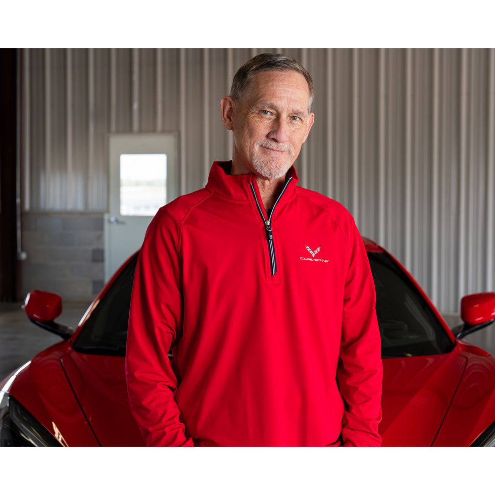 Man wearing a red quarter-zip jacket with a C7 Corvette logo standing next to a red Corvette in a garage.