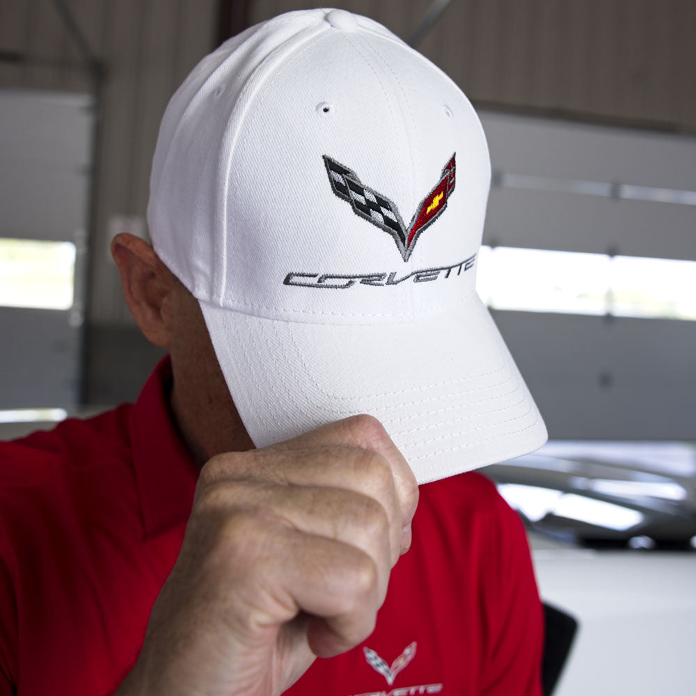 Man standing in a garage wearing a white baseball cap embroidered on the crown with a C7 Corvette emblem.