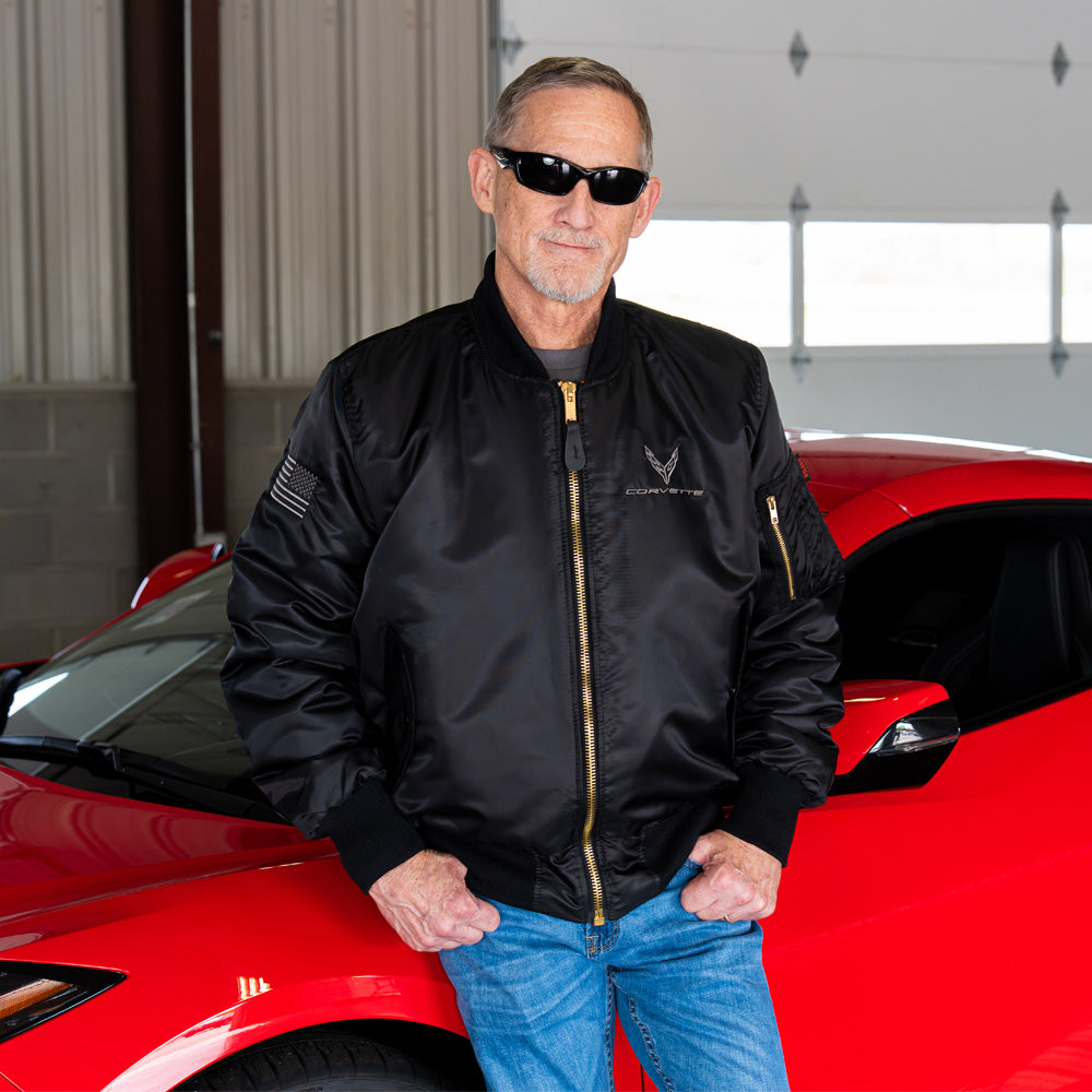 Man in a black aviator style jacket with an embroidered C8 Corvette emblem standing next to a red Corvette car in a garage.