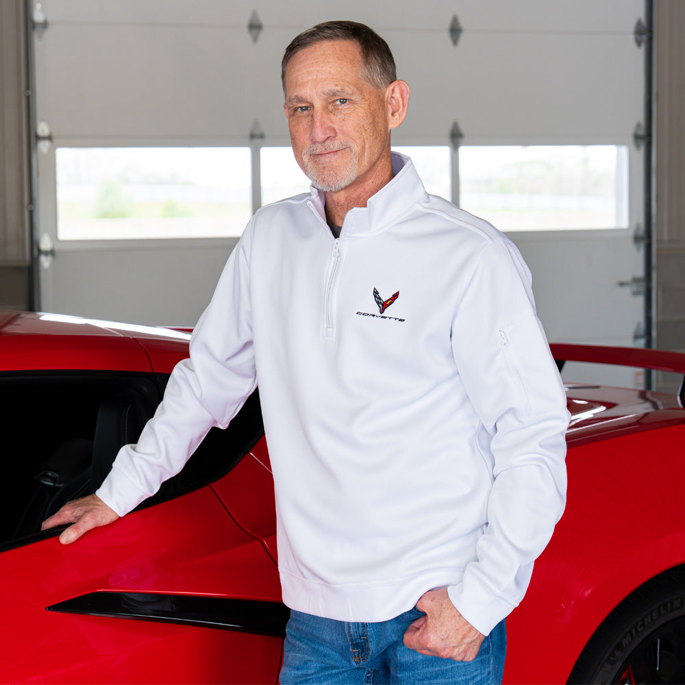 Man in a white quarter-zip pullover jacket with a C8 Corvette logo standing next to a red Corvette in a garage.