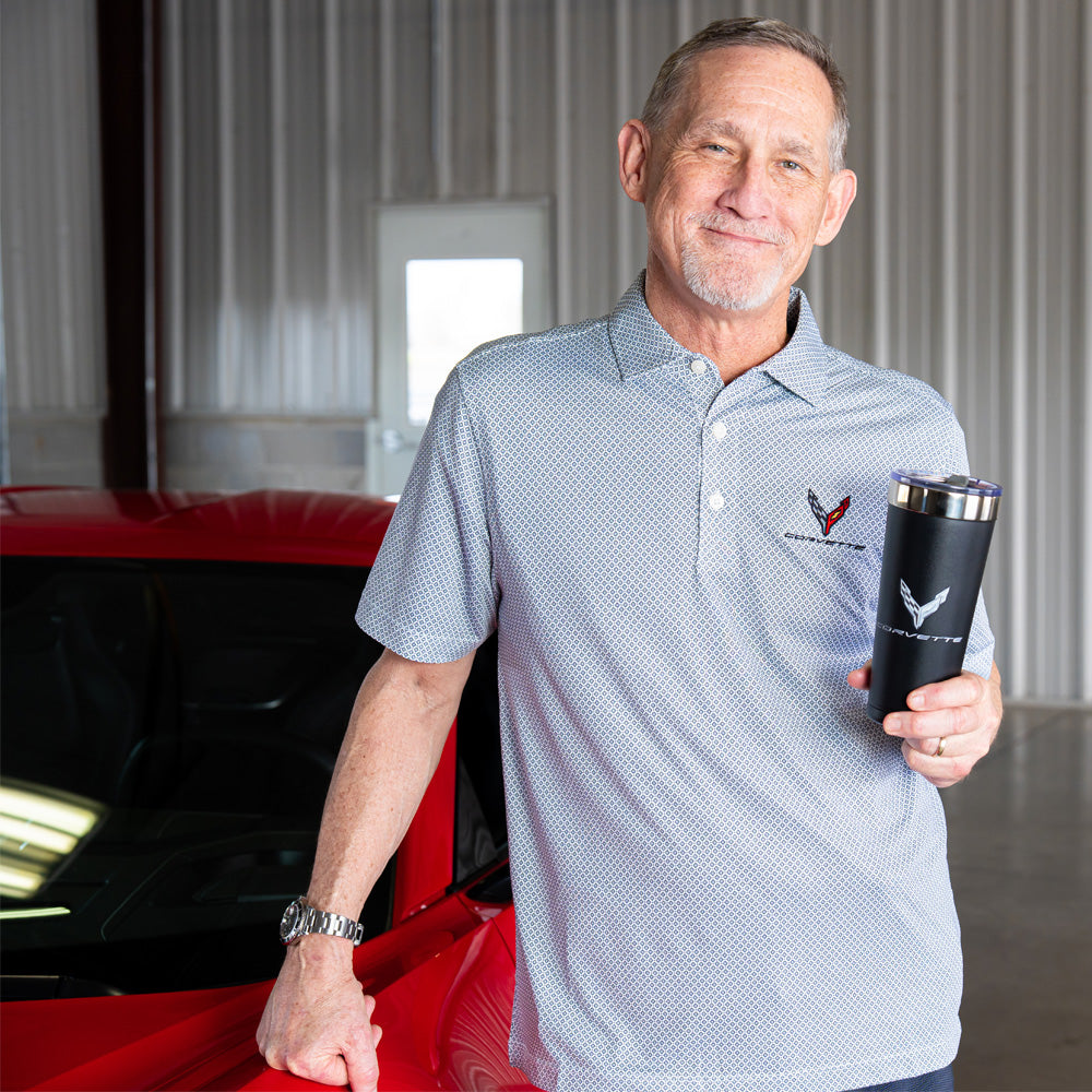 Man holding a black tumbler with a Corvette emblem and wearing the 8th Generation Corvette Emblem Diamond Dot Polo, standing next to a red Corvette in a garage.