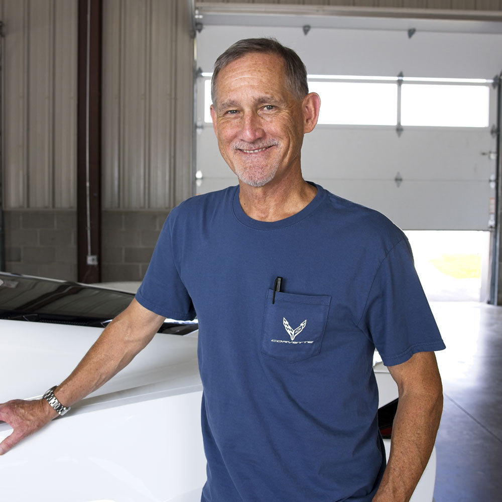 Man standing in a garage next to a white Corvette wearing a blue pocket tee with a C8 Corvette emblem on the pocket.
