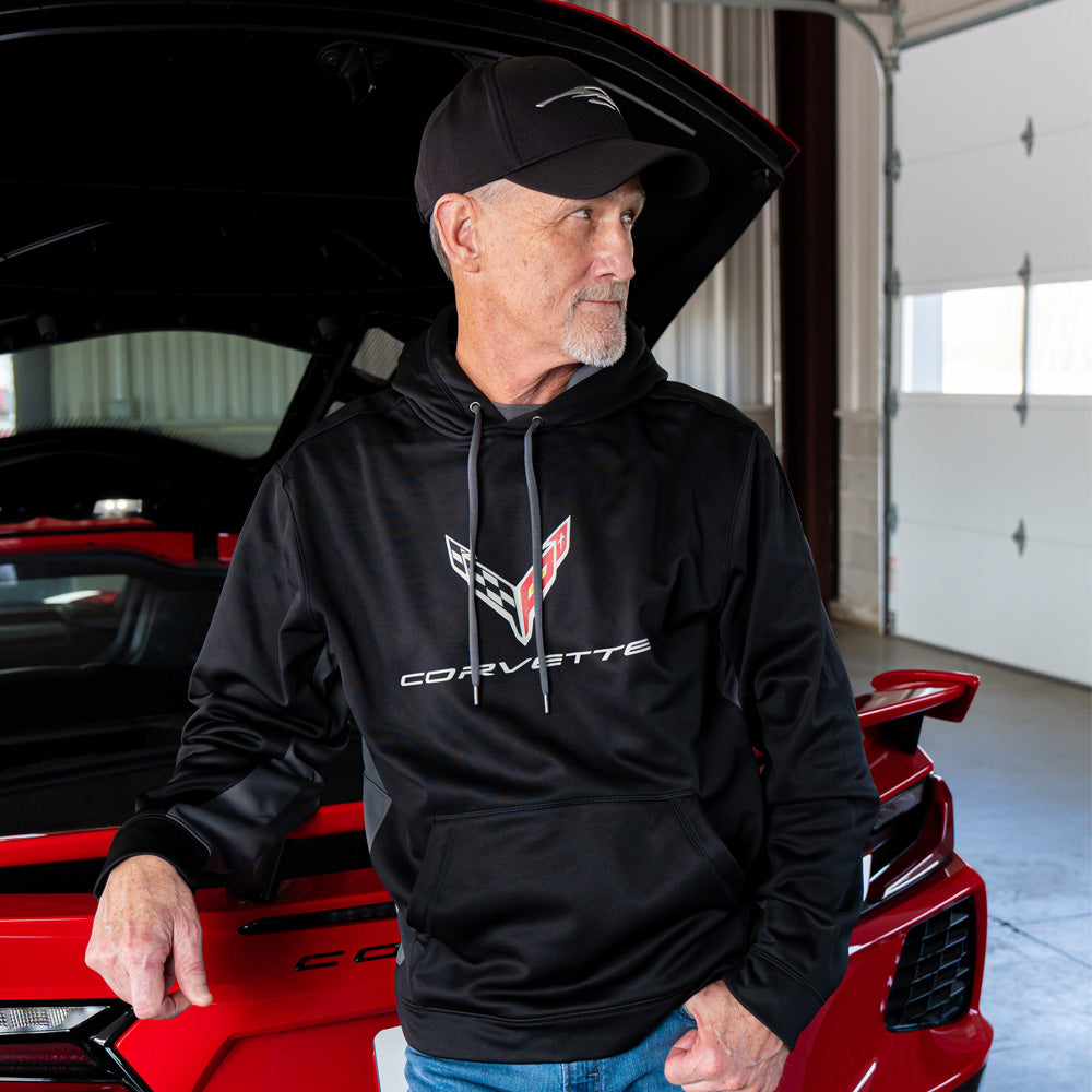 Man wearing a black Corvette hoodie standing next to a red Corvette in a garage.