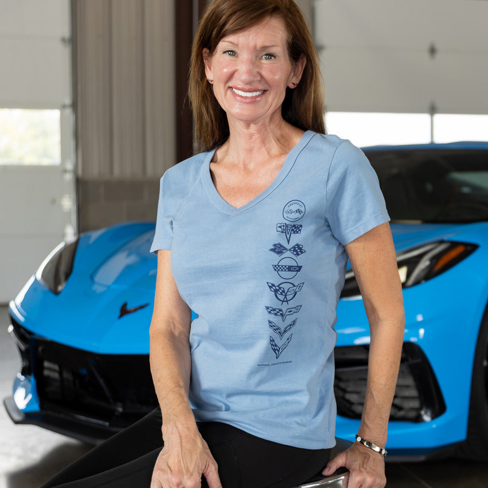 Woman wearing a blue t-shirt with a C1-C8 emblem design, standing in front of a blue Corvette.