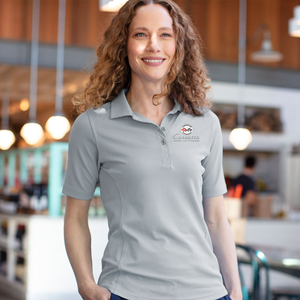 Woman standing in a cafe wearing the C1 Corvette Emblem Ladies Core Polished Gray Virtue Polo