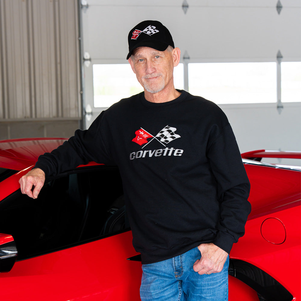 Man wearing a black Corvette sweatshirt and cap standing next to a red Corvette in a garage.