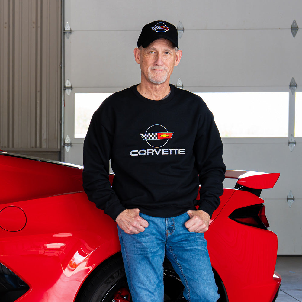 Man wearing a Corvette sweatshirt with a C4 Corvette emblem standing next to a red Corvette in a garage.