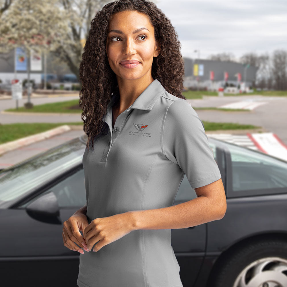 Woman wearing the C5 Corvette Emblem Ladies Core Polished Gray Virtue Polo while standing in front of a black C5 Corvette