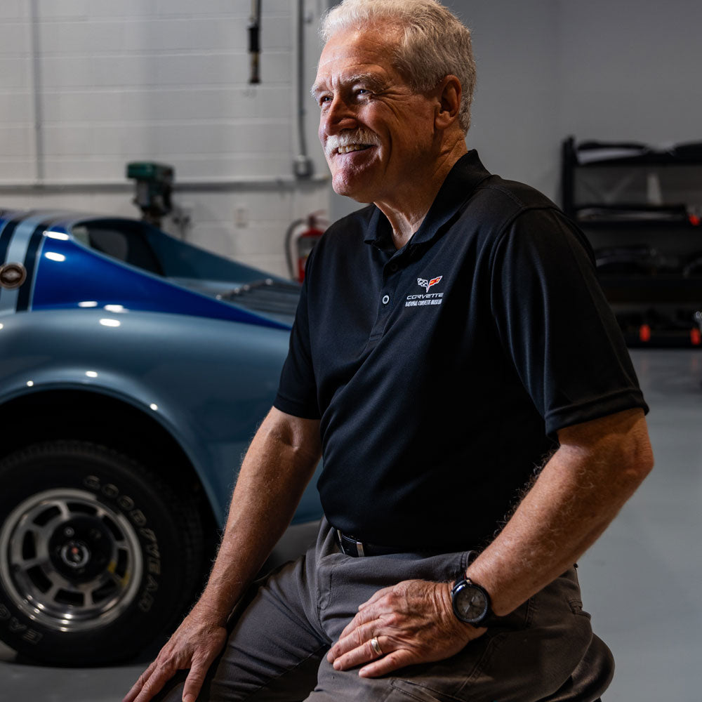 Man sitting on a stool in a garage wearing a black C6 Corvette Polo