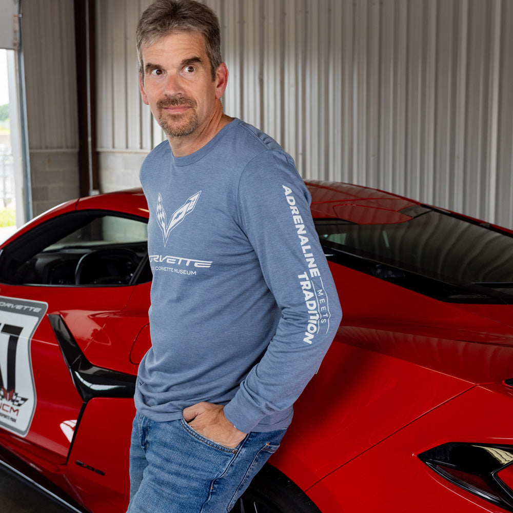 Man in a blue Corvette long sleeve t-shirt standing next to a red Corvette in a garage.