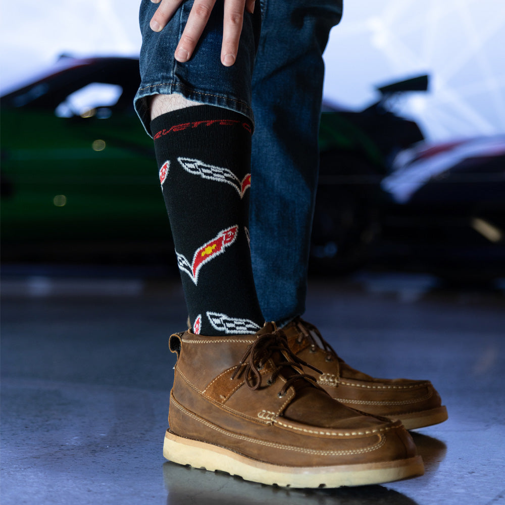 Person wearing black socks decorated with C7 Corvette emblems and brown boots in a blurred indoor setting.