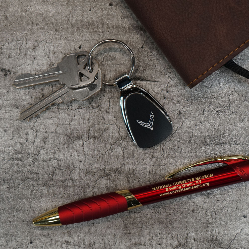 C8 Corvette Emblem Chrome Teardrop Keychain Displayed on a table with a notebook and pen