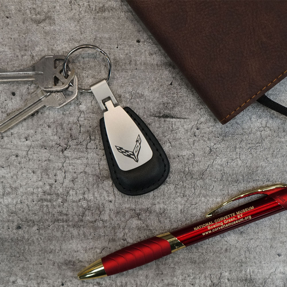 The C8 Corvette Emblem Leather Teardrop Keychain shown on a table next to a pen and notebook