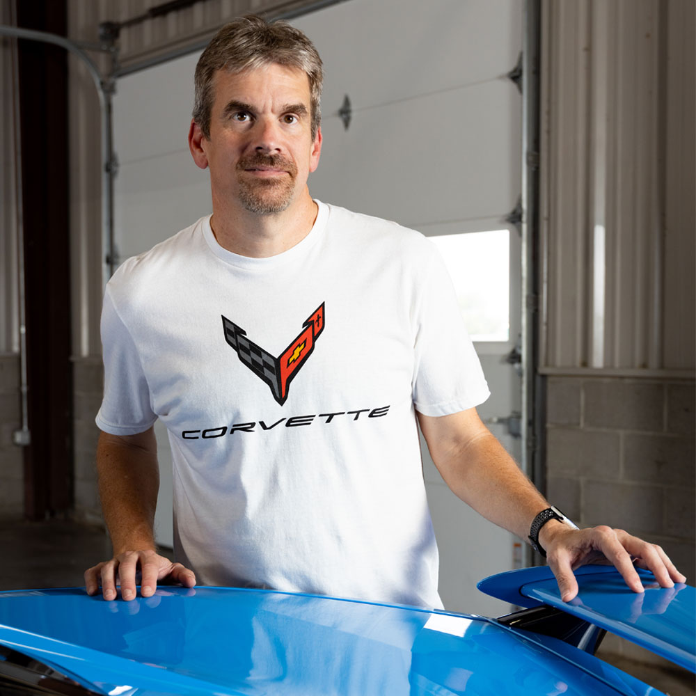 A man wearing a white C8 Corvette t-shirt stands behind a bright blue Corvette in a garage setting.