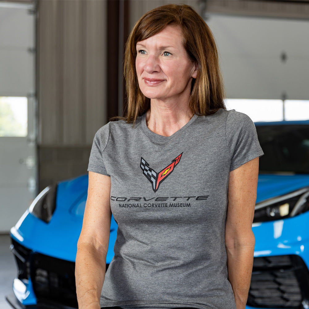 Woman wearing a gray C8 Corvette tee shirt standing in front of a blue Corvette in a garage.
