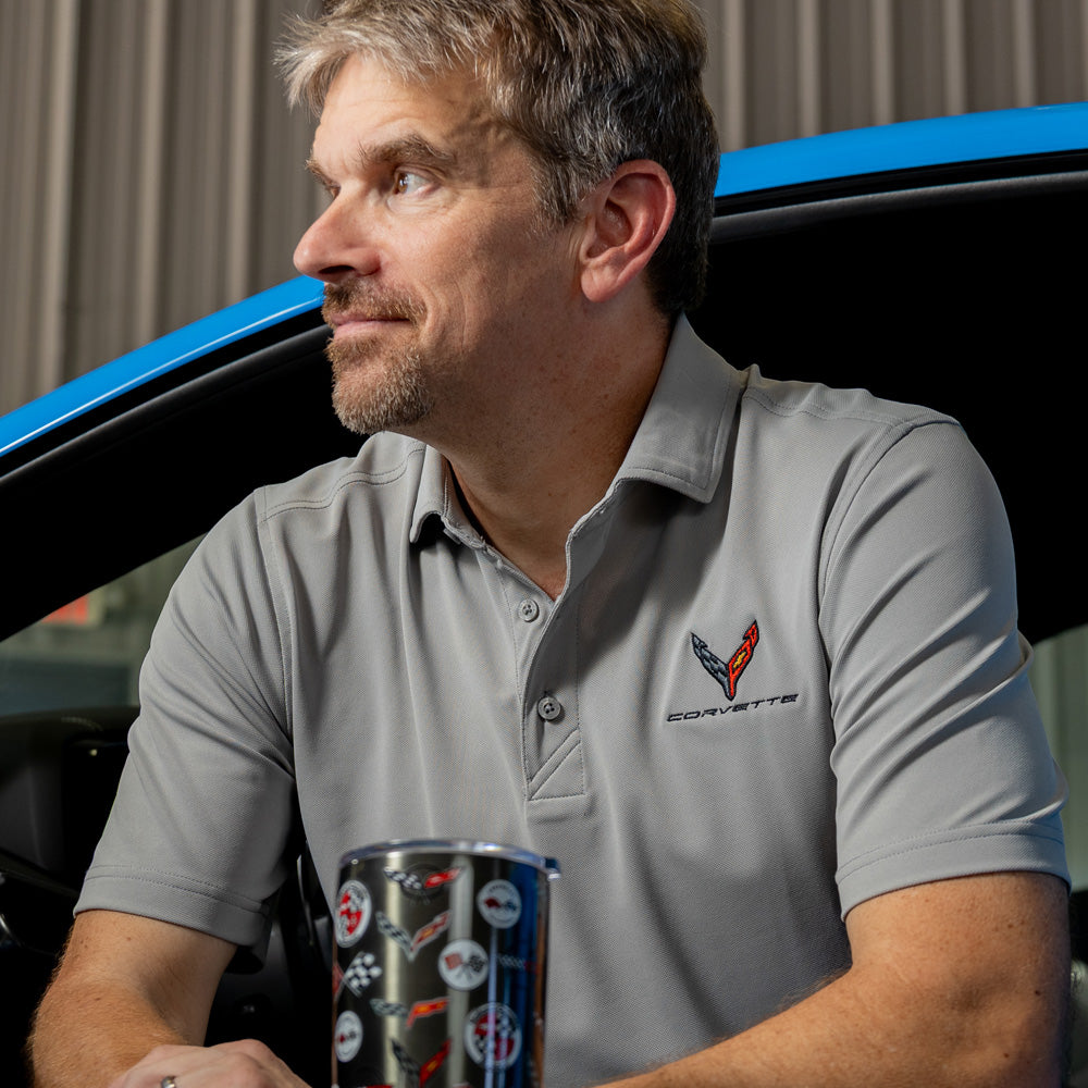Man wearing a gray C8 Corvette polo shirt holding a travel mug in front of a blue Corvette.