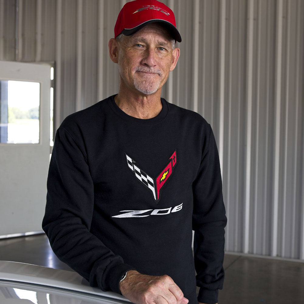 A man standing next to a white Corvette wearing a black crewneck sweatshirt with an embroidered C8 Z06 Corvette emblem on the front. 