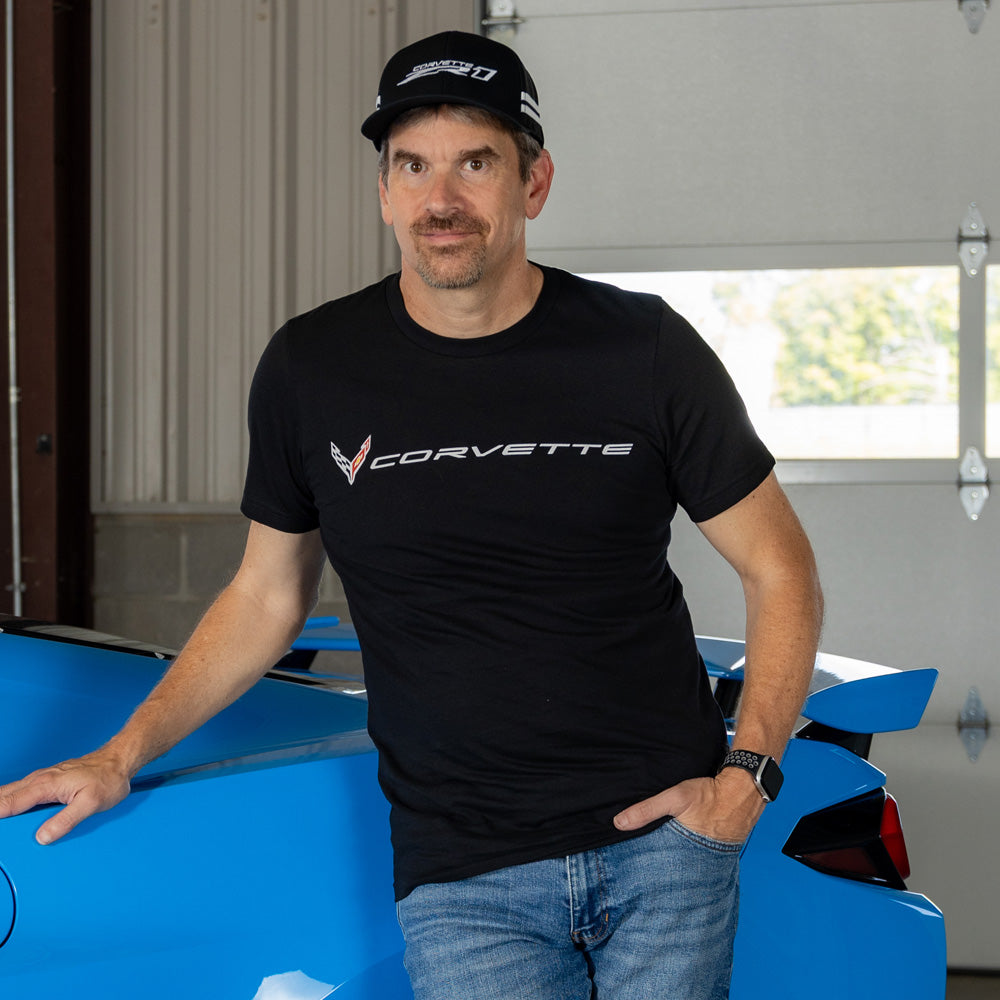 Man wearing a black C8 ZR1 Corvette t-shirt standing next to a blue Corvette in a garage.