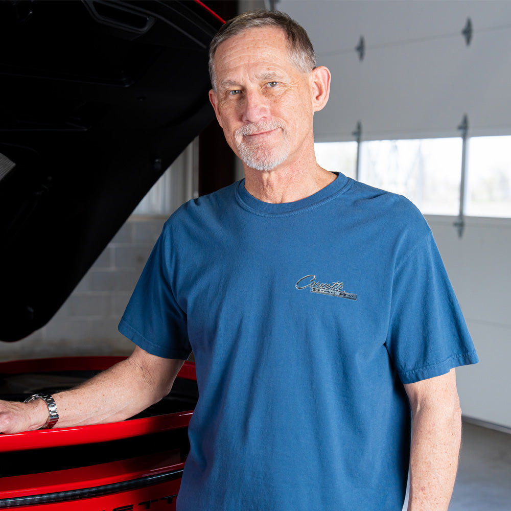 Man wearing a blue t-shirt with a Corvette Sting Raylogo, standing next to a red Corvette in a garage.
