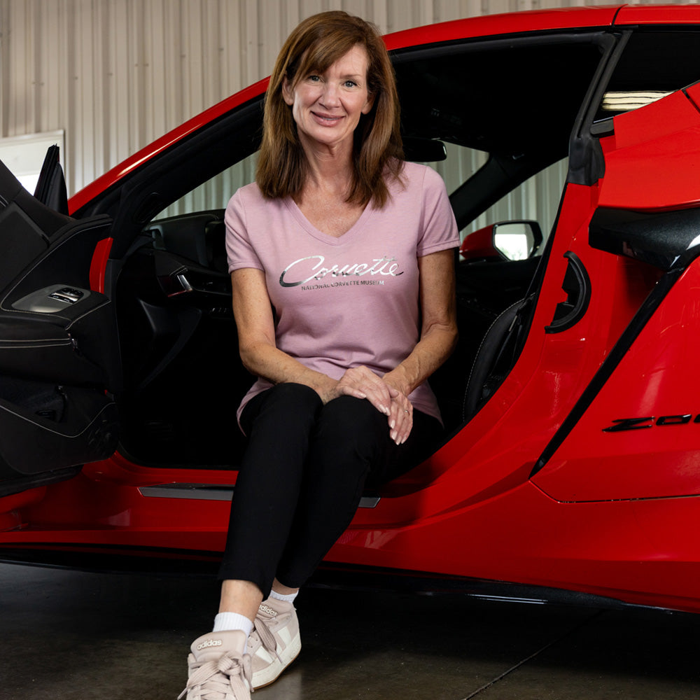 Woman sitting inside a red Corvette with open door wearing the Corvette Foil Script Ladies Mauve tee.