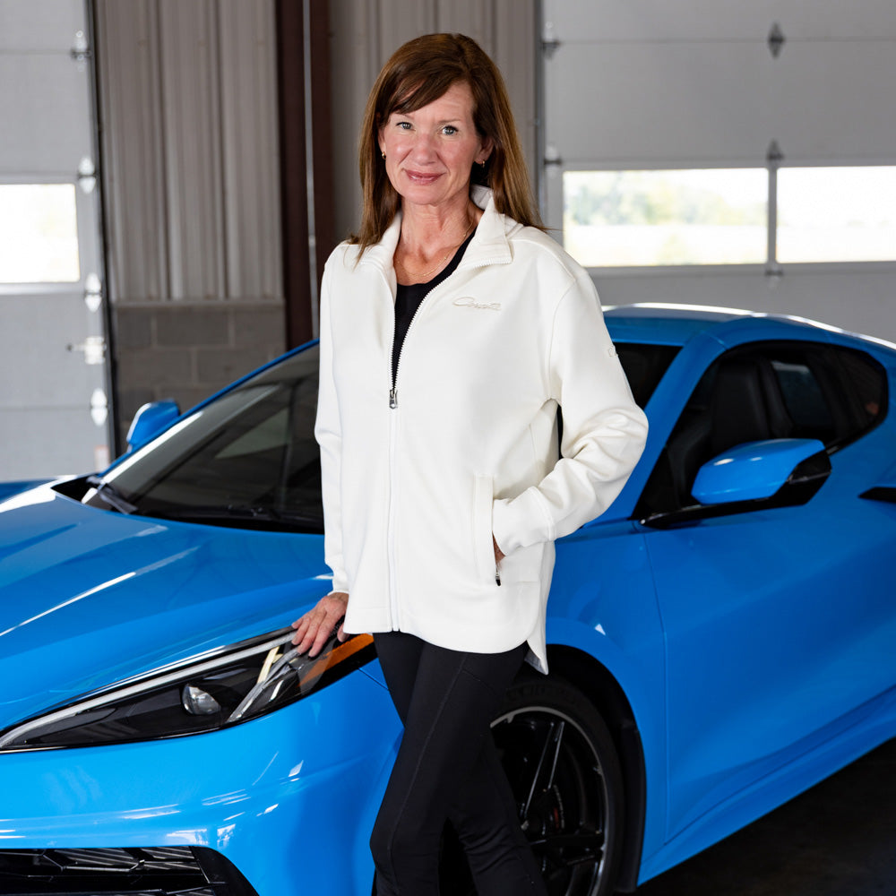 Woman in a white jacket with Corvette script standing next to a blue Corvette in a garage.