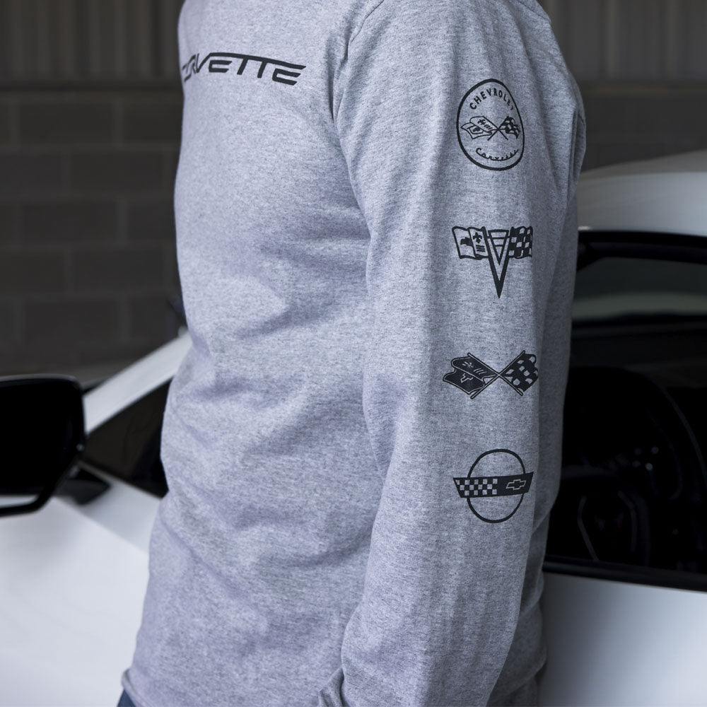 A man standing next to a white Corvette wearing a long sleeve gray tee showing a close up of the C1-C4 Corvette emblems on the left sleeve.