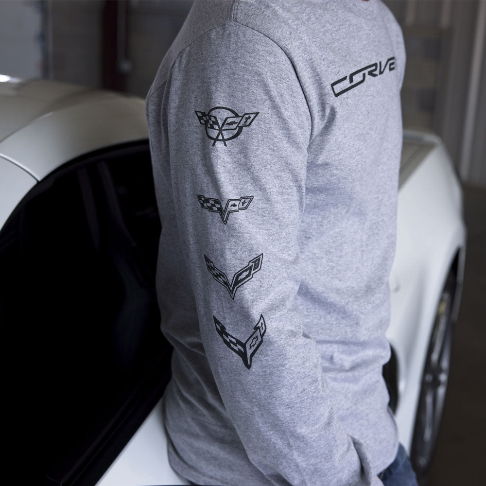 A man standing next to a white Corvette wearing a long sleeve gray tee with the C5 - C8 Generation Corvette emblems showing printed down the right sleeve.