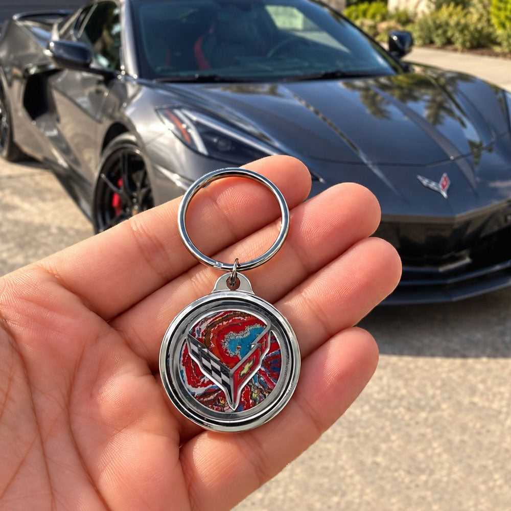 A person holding the Corvette Paint 8th Generation Corvette Emblem Keychain in their hand in front of a black C8 Corvette.
