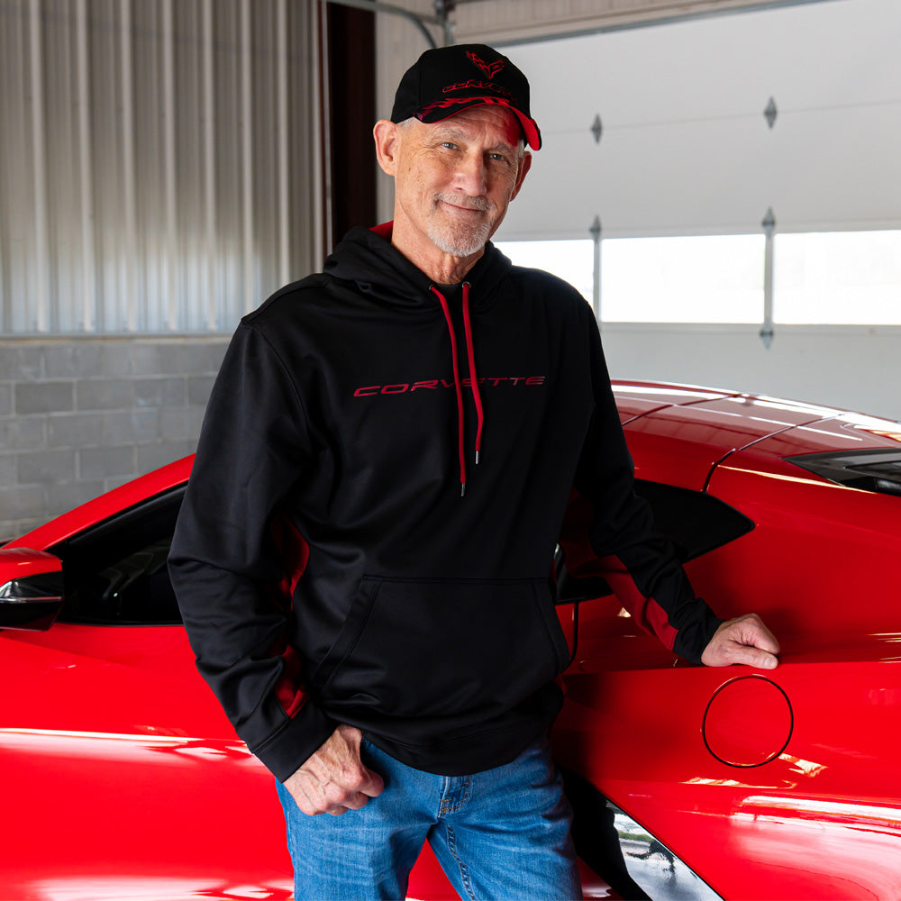 Man standing in a garage next to a red Corvette wearing the Corvette Script Sport-Tek Colorblock Black and Red Hoodie 