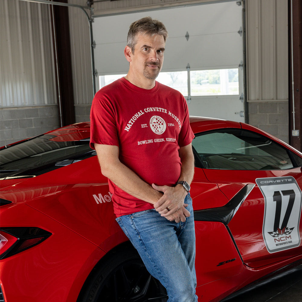 A man wearing a red t-shirt with a 'National Corvette Museum Bowling Green Arena Center' graphic and blue jeans leans against a red Corvette sports car inside a garage.